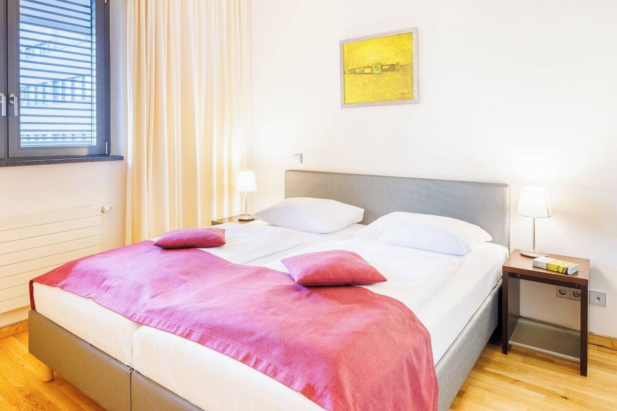 A hotel room with a double bed, two bedside tables with lamps, a window with blinds, and a framed artwork on the wall.