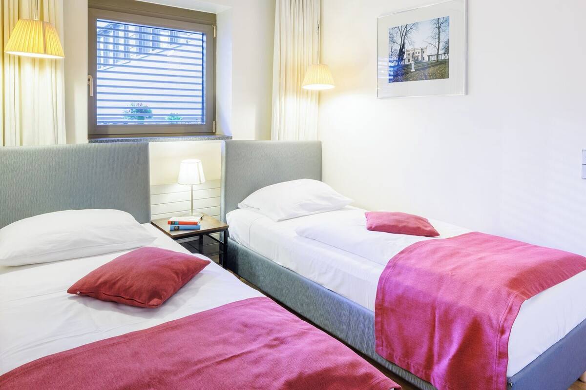 A hotel room with two single beds, a nightstand with a lamp, a window with blinds, and a framed picture on the wall.
