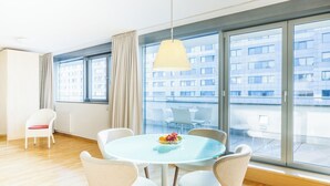 Una habitación moderna con una mesa redonda de vidrio, cuatro sillas, una lámpara colgante y grandes ventanas que ofrecen vista a un edificio.