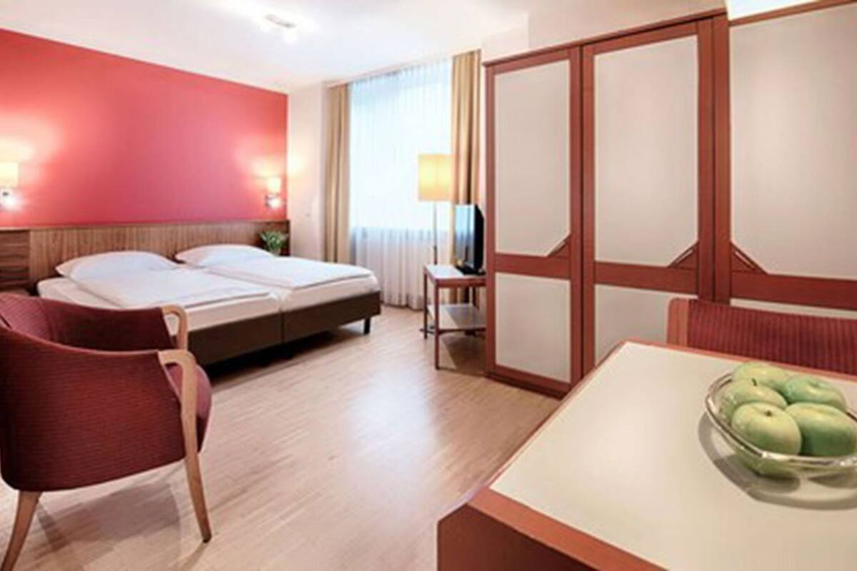 A hotel room with a bed, a chair, a table with apples, and a TV.