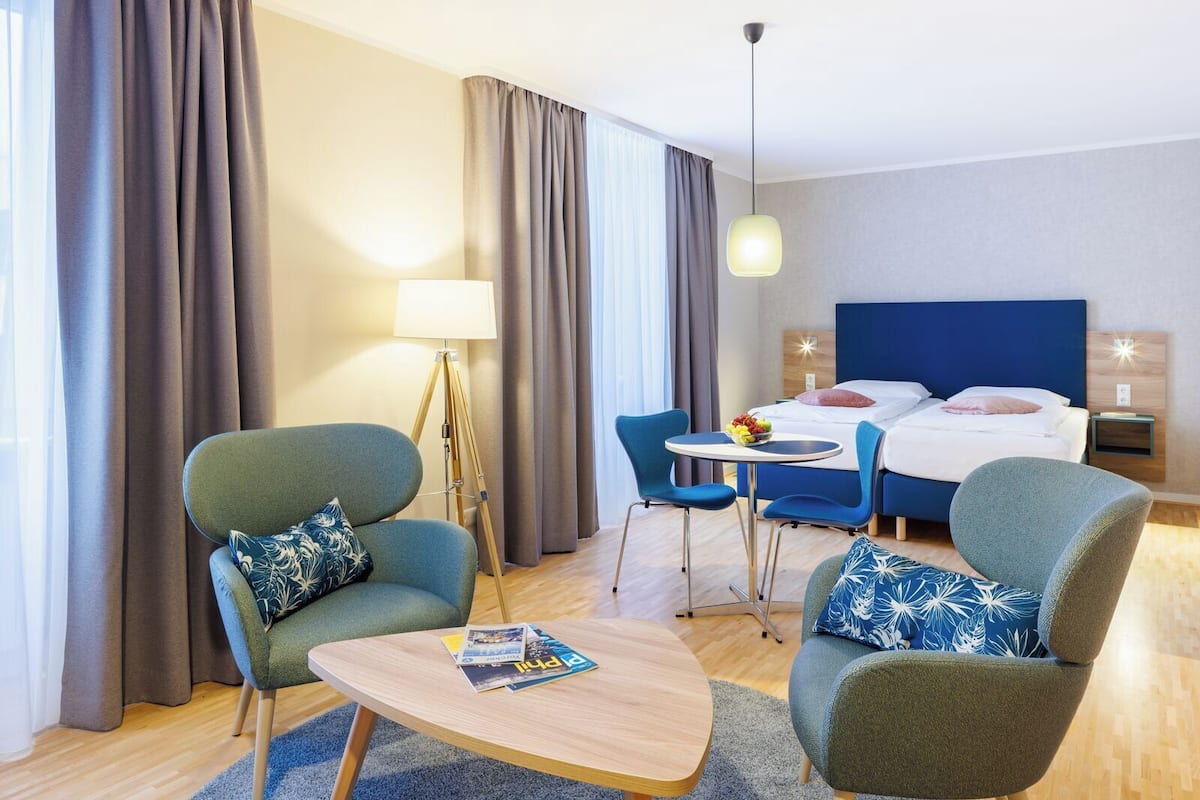 A hotel room with a bed, two armchairs, a small table, a lamp, and a dining table with chairs.