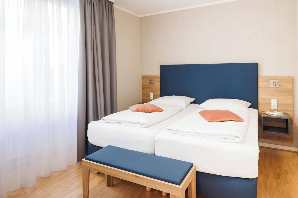 A double bed with two pillows, a bedside table with a book, a wooden bench, and a window with curtains.