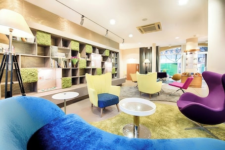 A modern lounge area with a sofa, yellow and purple chairs, a small table, and a bookshelf with green plants.