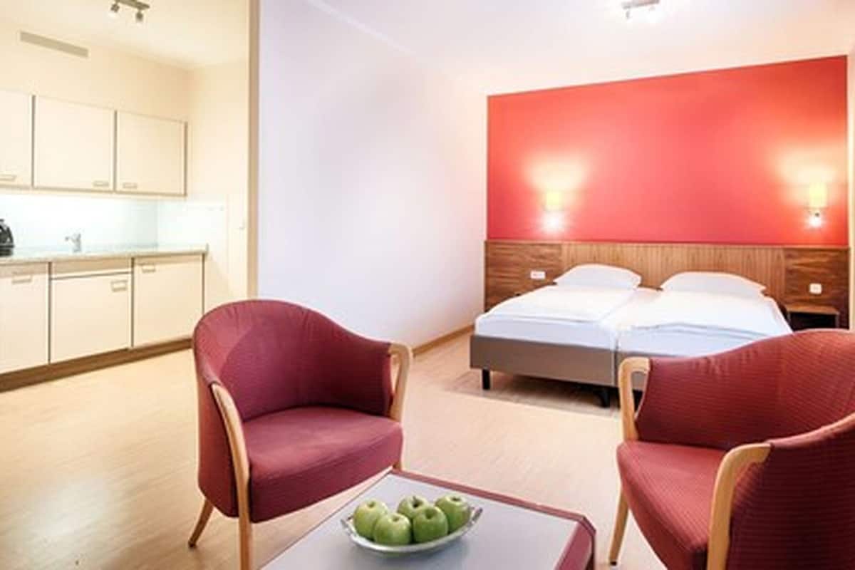 A hotel room with a bed, two red chairs, a small table with apples, and a kitchen area.