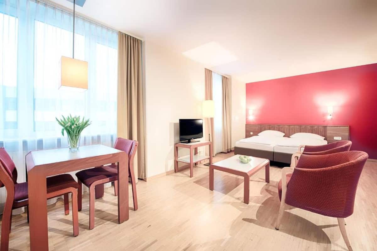 A hotel room with a bed, a TV, a dining table with chairs, a coffee table, and a plant.