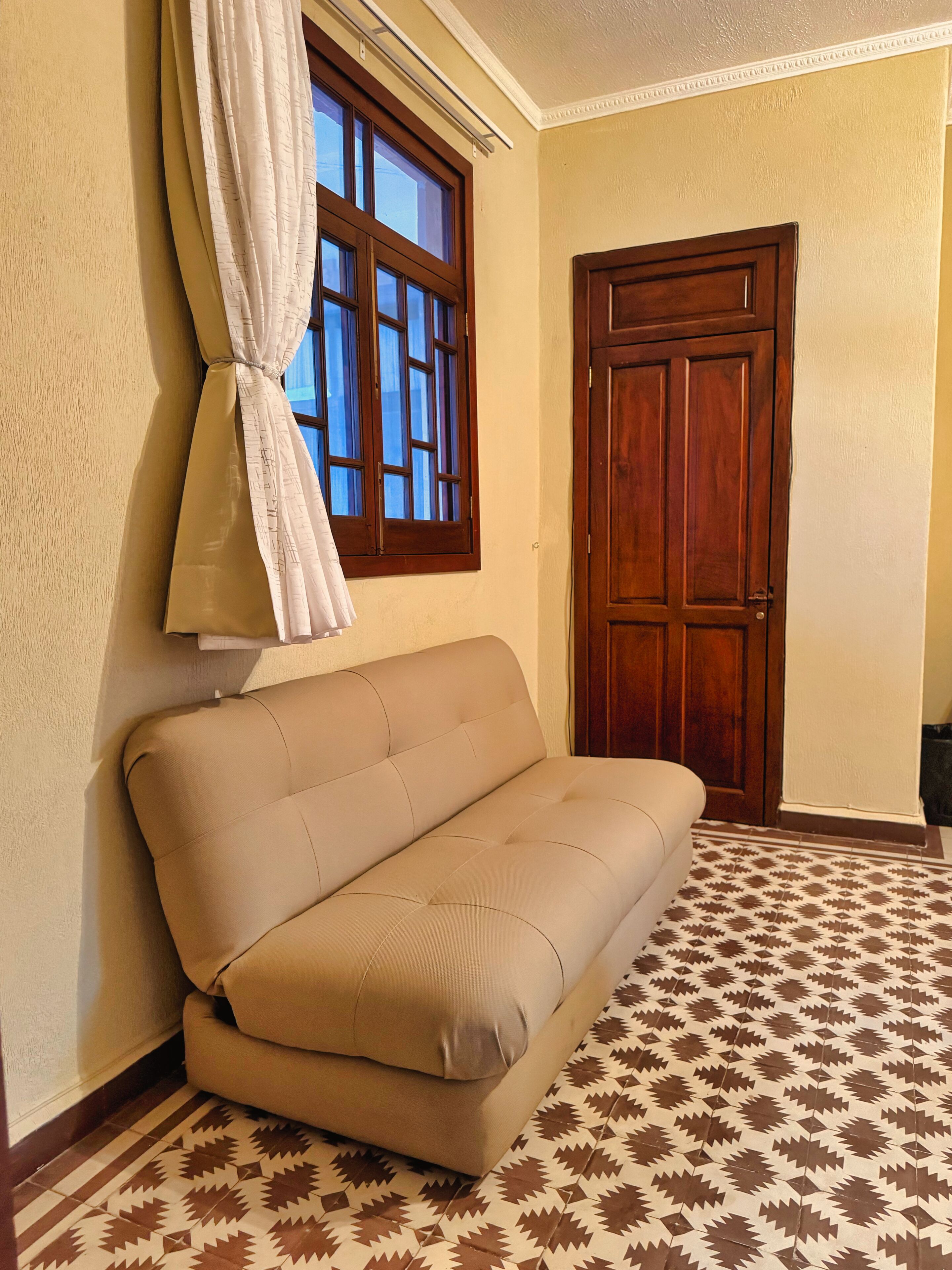 A room with a sofa against a wall, a window with curtains, a wooden door, and patterned floor tiles.