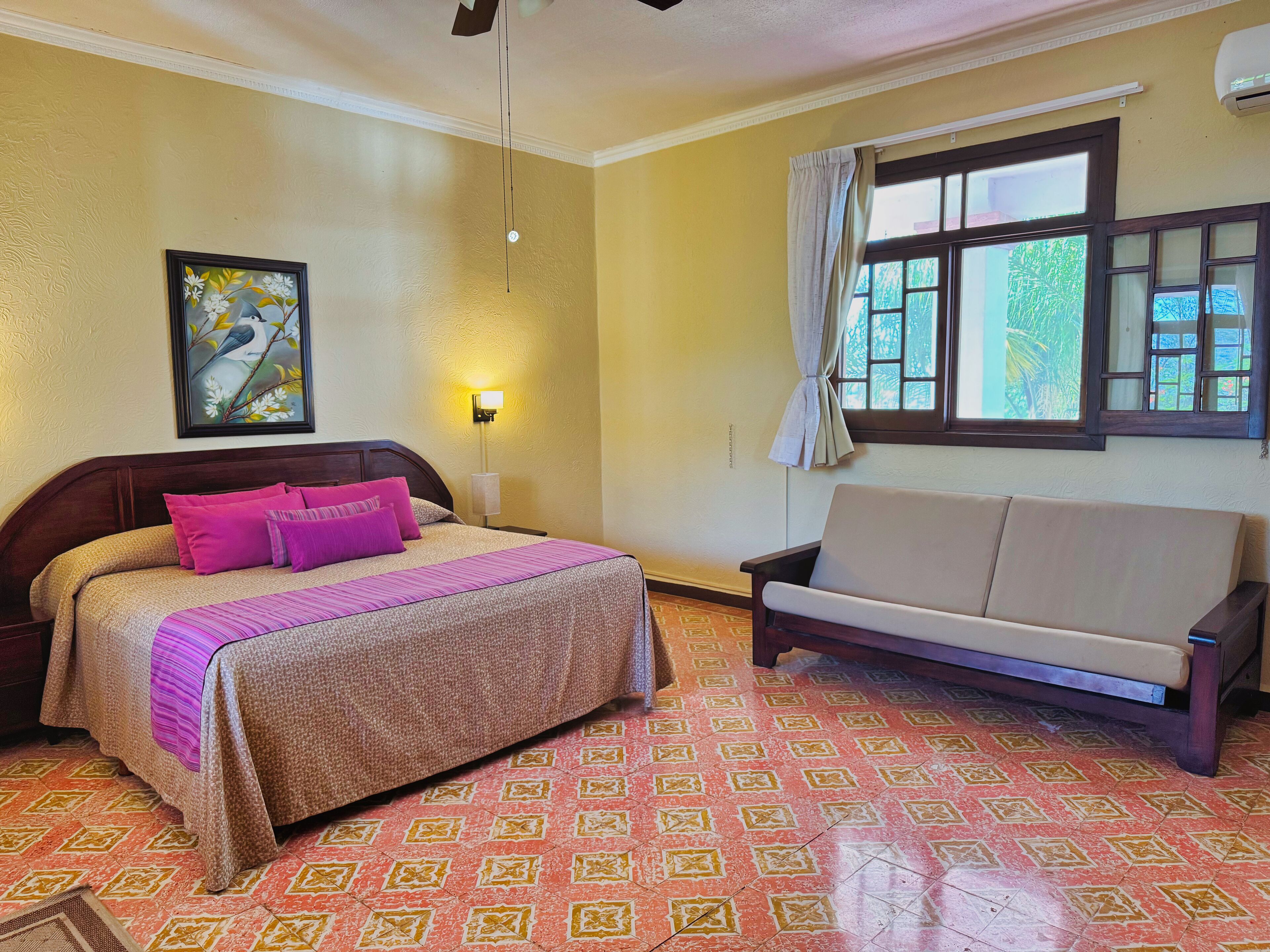 Una habitación de hotel con cama, sofá, un cuadro, ventana con cortinas y suelo estampado.