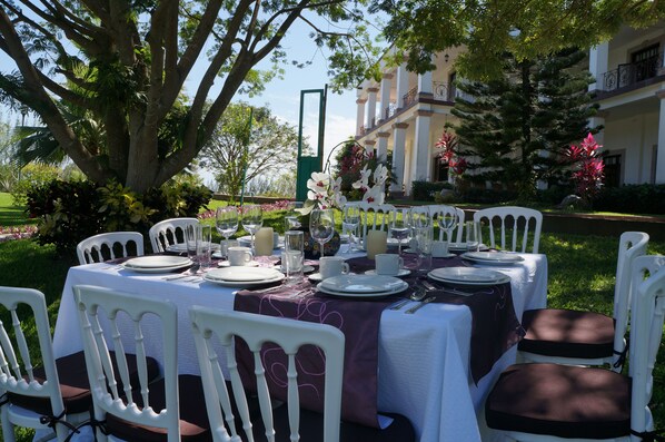 An outdoor dining setup with white chairs and tables, surrounded by lush greenery and a large tree.