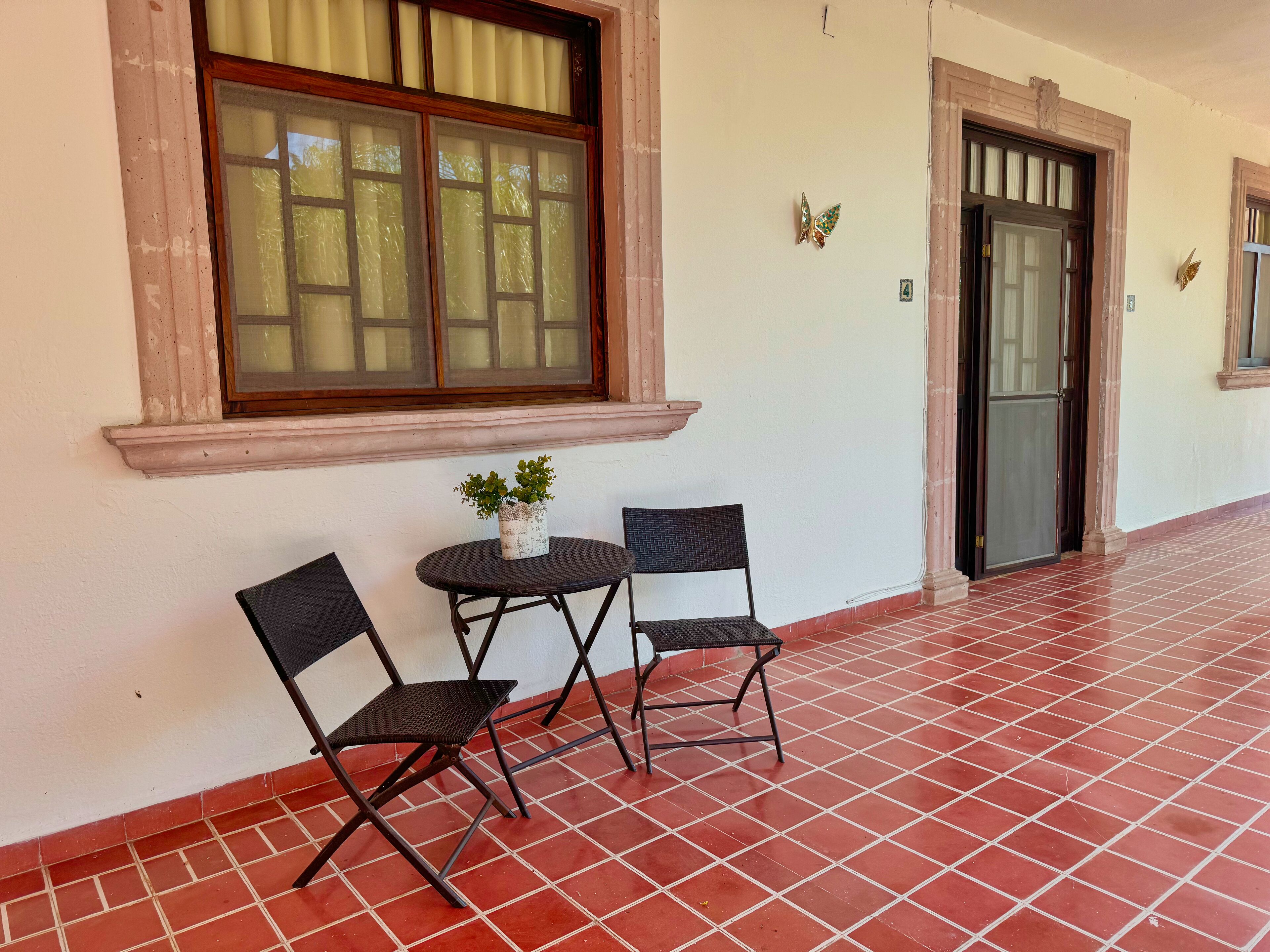 A corridor with a tiled floor, a window with curtains, two chairs, a small table with a plant, and a door with a number plate.