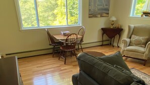 A living room with a wooden floor, a gray sofa, a dining table with chairs, and two windows with a view of trees. - Quintessential Cape Vibes in Boothbay (Boothbay)