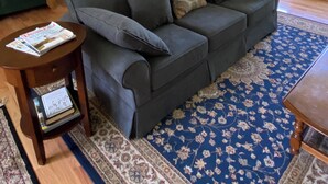 A living room with a grey sofa, a blue patterned rug, a wooden side table with magazines, a staircase with wooden railings, and a dining area with a wooden table and chairs. - Quintessential Cape Vibes in Boothbay (Boothbay)