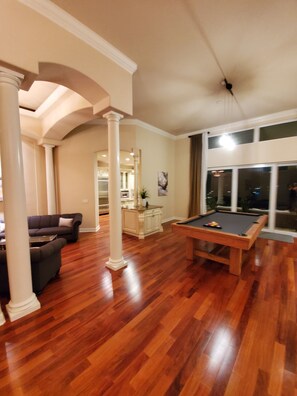A spacious living room with a pool table, a sofa, and a kitchen area in the background. - Luxury Pool Spa Home with Spectacular Water View! (Cape Coral)