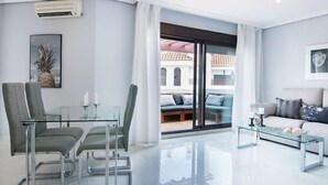 Ein modernes Wohnzimmer mit einem Glastisch, zwei Stühlen, einem Sofa und einem Couchtisch. Ein großes Schiebe-Glasfenster führt auf eine Terrasse.