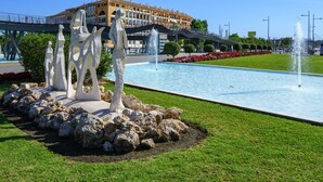 Ein Springbrunnen mit Becken, umgeben von Steinen und Gras, mit einem Gebäude im Hintergrund.