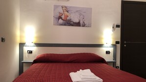 A neatly made bed with a red bedspread and white towels, two bedside lamps, a painting on the wall, and a door to the right. - La Casa di Anna (Delebio)