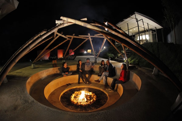 A group of people sitting around a circular fire pit under a metal arch structure at night.