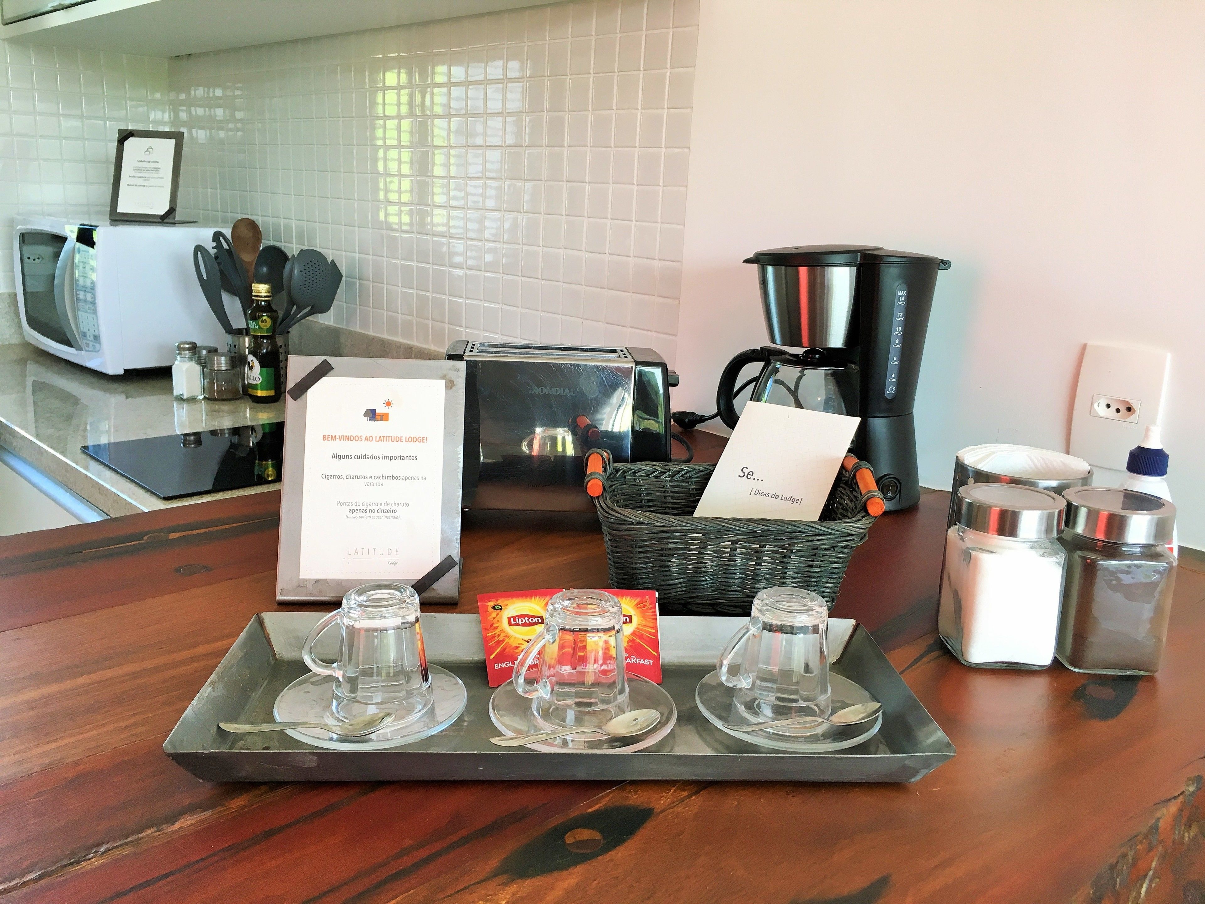 A kitchen counter with a tray of clear glass mugs, a toaster, a coffee maker, salt and pepper shakers, and a sign with hotel information.