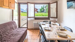 Uma área de refeições com mesa posta para quatro pessoas, um sofá e vista para uma paisagem verde através de janelas abertas.
