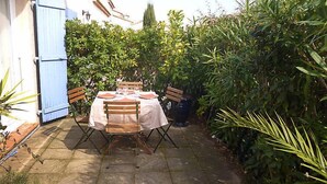 Une terrasse extérieure avec une table dressée pour quatre personnes, entourée d’une végétation luxuriante et d’un bâtiment blanc aux volets bleus.