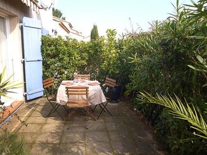 Eine Terrasse im Freien mit Tisch für vier Personen, umgeben von üppiger Vegetation und einem weißen Gebäude mit blauen Fensterläden.
