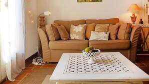 Un salon avec un canapé beige, une table basse blanche sur laquelle se trouve un panier de fruits, et un tableau représentant un paysage accroché au mur.