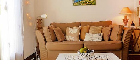 Un salon avec un canapé beige, une table basse blanche sur laquelle se trouve un panier de fruits, et un tableau représentant un paysage accroché au mur.