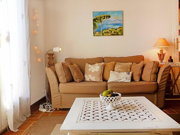 A living room with a beige sofa, a white coffee table with a fruit bowl, and a painting of a landscape on the wall.