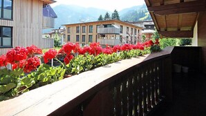 Een balkon met rode bloemen en houten hekwerk met uitzicht op een woongebied met moderne gebouwen en bergen op de achtergrond.