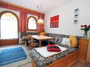 Una habitación acogedora con un banco de madera, una mesa de comedor y sillas. Hay dos ventanas con cortinas, una alfombra en el suelo y una pared con cuadros.
