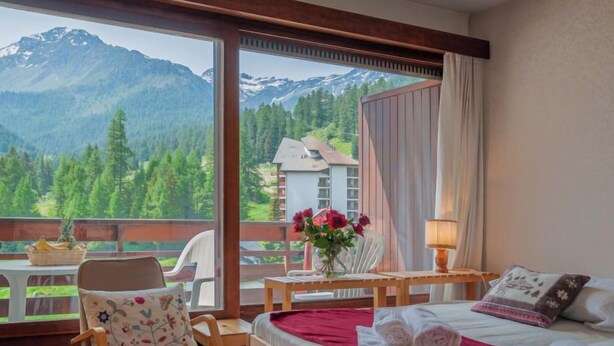 A hotel room with a bed, a chair, a table with a vase of flowers, and a view of mountains and trees through large windows. - Rosablanche B72 by Interhome (Siviez-Nendaz)