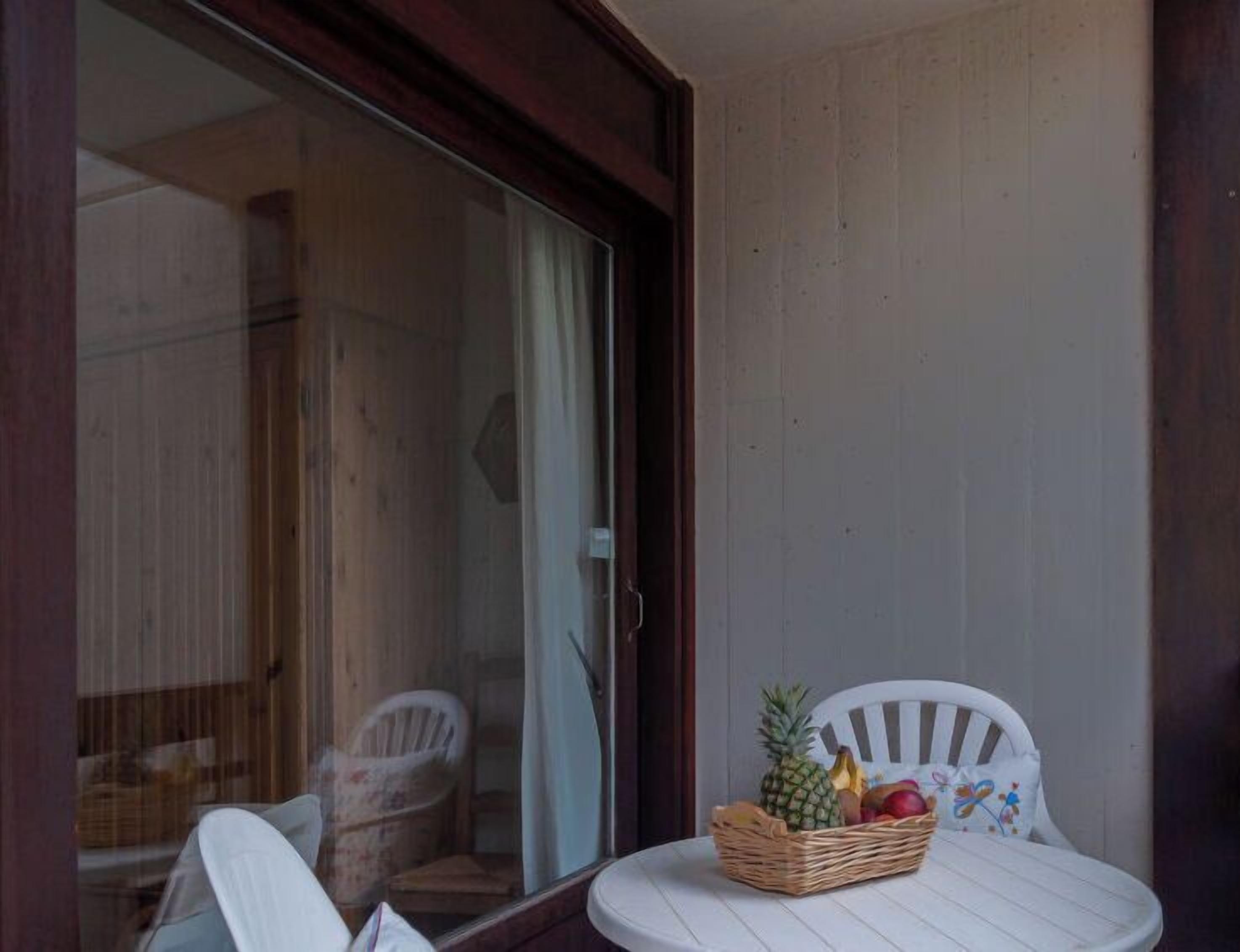 A balcony with a white chair, a wicker fruit basket, and a sliding glass door leading to an interior space with a wooden floor and a white chair.