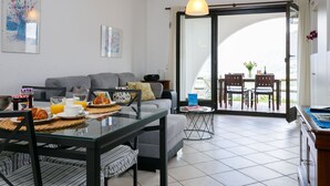 Un salon moderne avec une table à manger en verre, un canapé gris et une vue sur un balcon aménagé avec du mobilier d’extérieur.