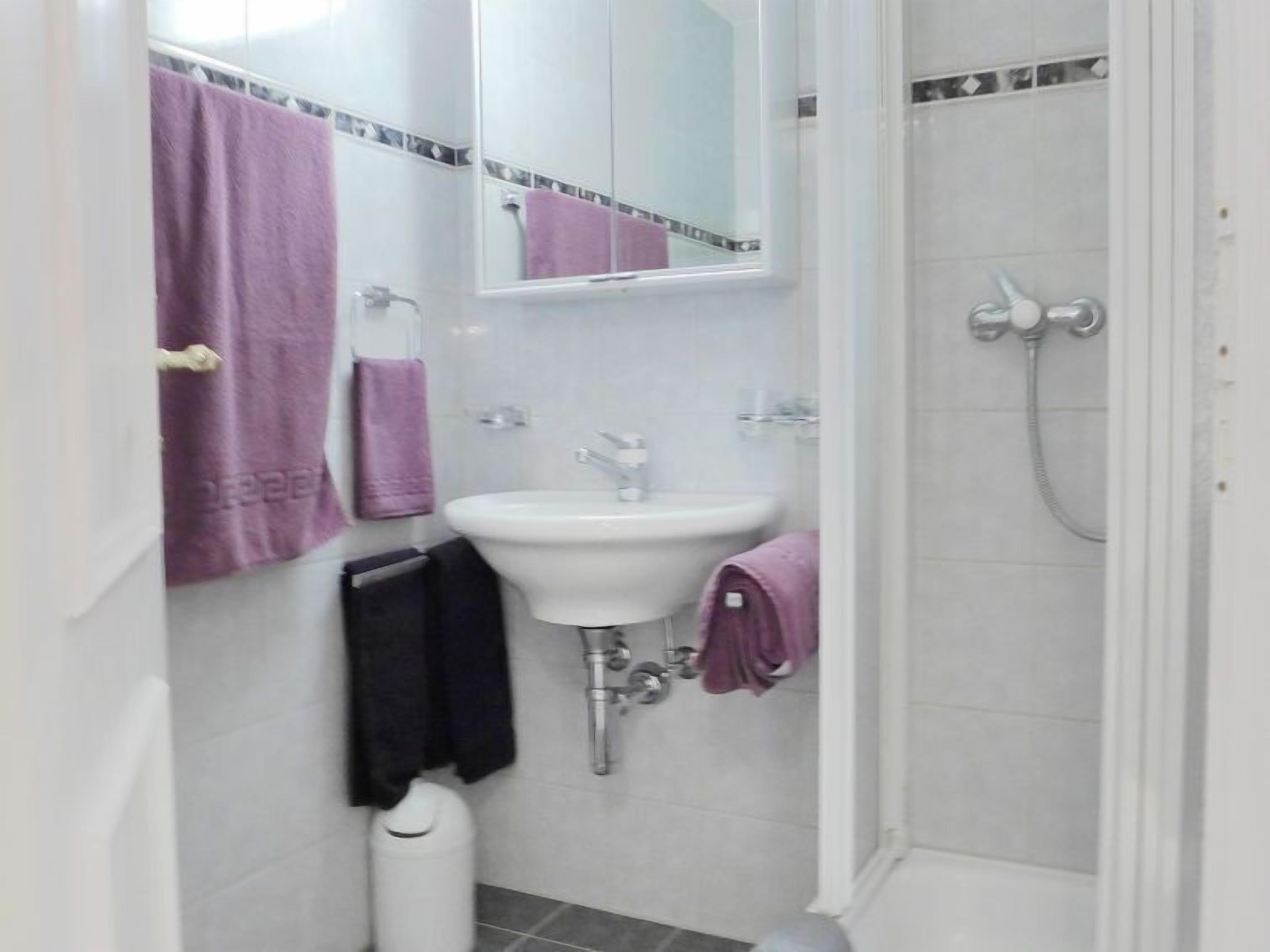 Un baño pequeño con un lavabo blanco, un espejo y una ducha. Toallas púrpuras cuelgan de la pared.