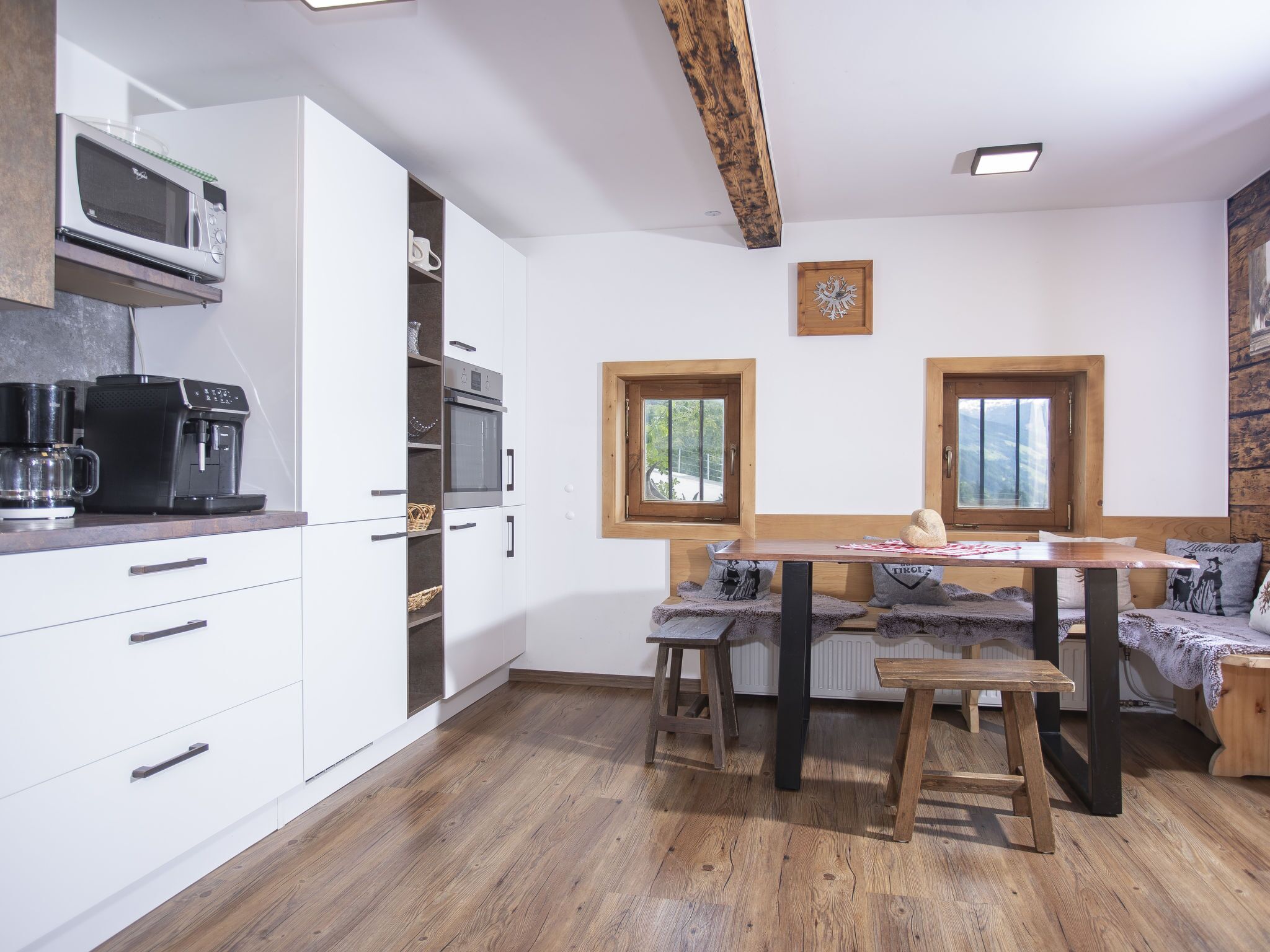 Eine moderne Küche mit weißen Schränken, integriertem Mikrowellenherd, einer Kaffeemaschine und einem Holztisch mit Barhocker. Zwei Fenster mit Holzrahmen bieten Blick ins Grüne.