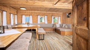 A wooden cabin interior with a dining table, benches, a sofa, and a patterned carpet.