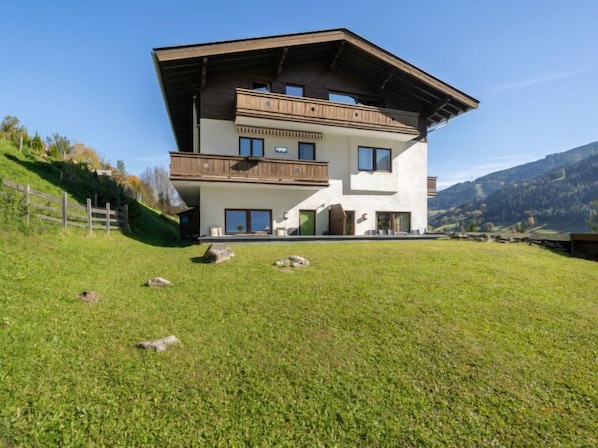 Uma casa moderna de dois andares com telhado inclinado e varanda de madeira, com montanhas ao fundo.