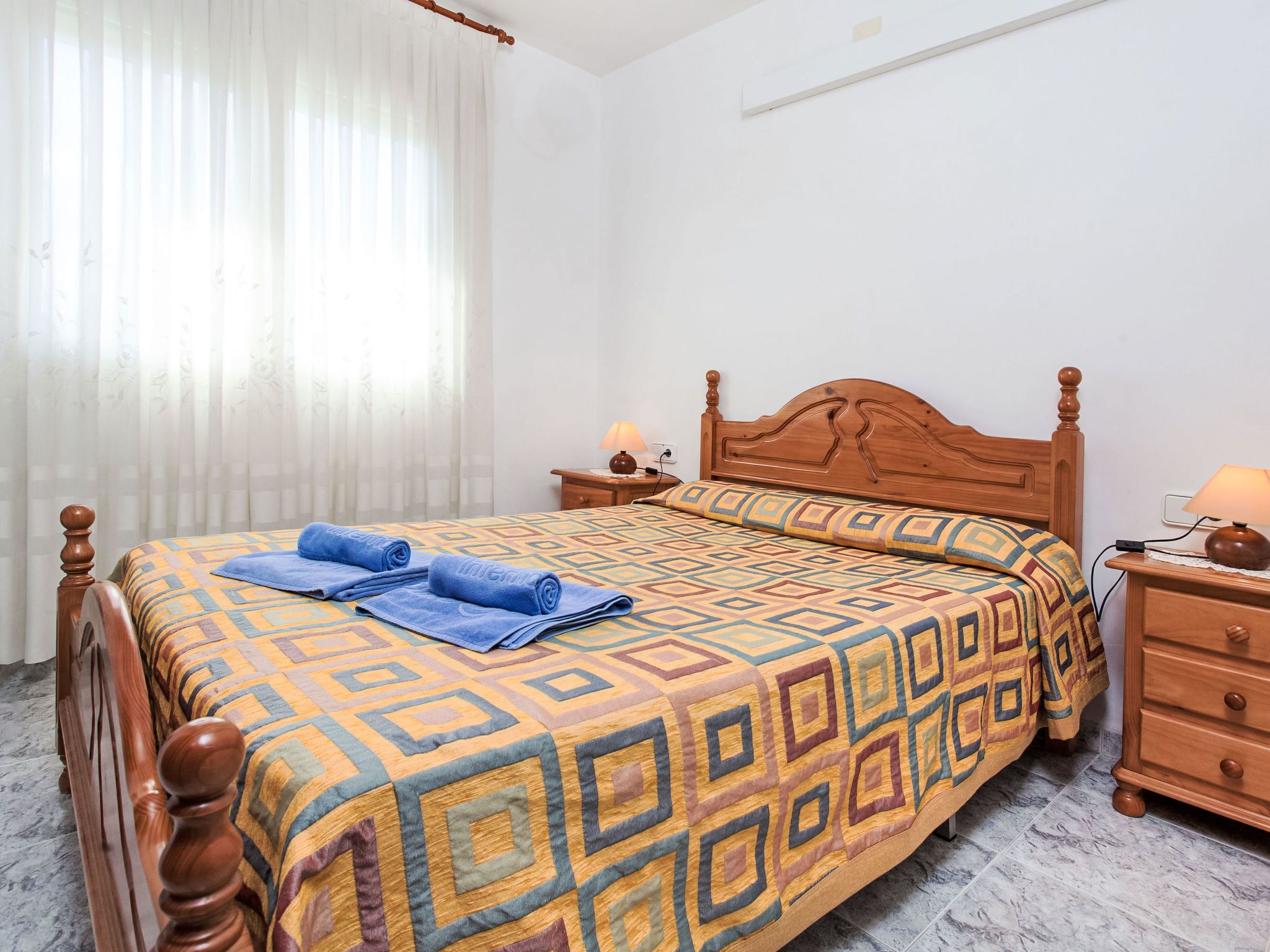 Una cama bien hecha con colcha estampada y toallas enrolladas, una mesita de noche de madera con lámpara, y una ventana con cortinas traslúcidas.