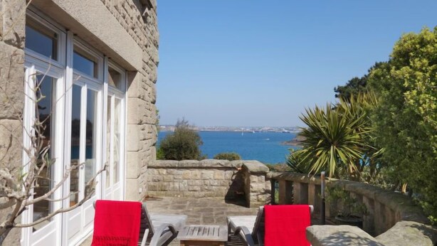 Edificio de piedra con terraza que presenta dos sillones con cojines rojos, una mesita y vistas al mar.