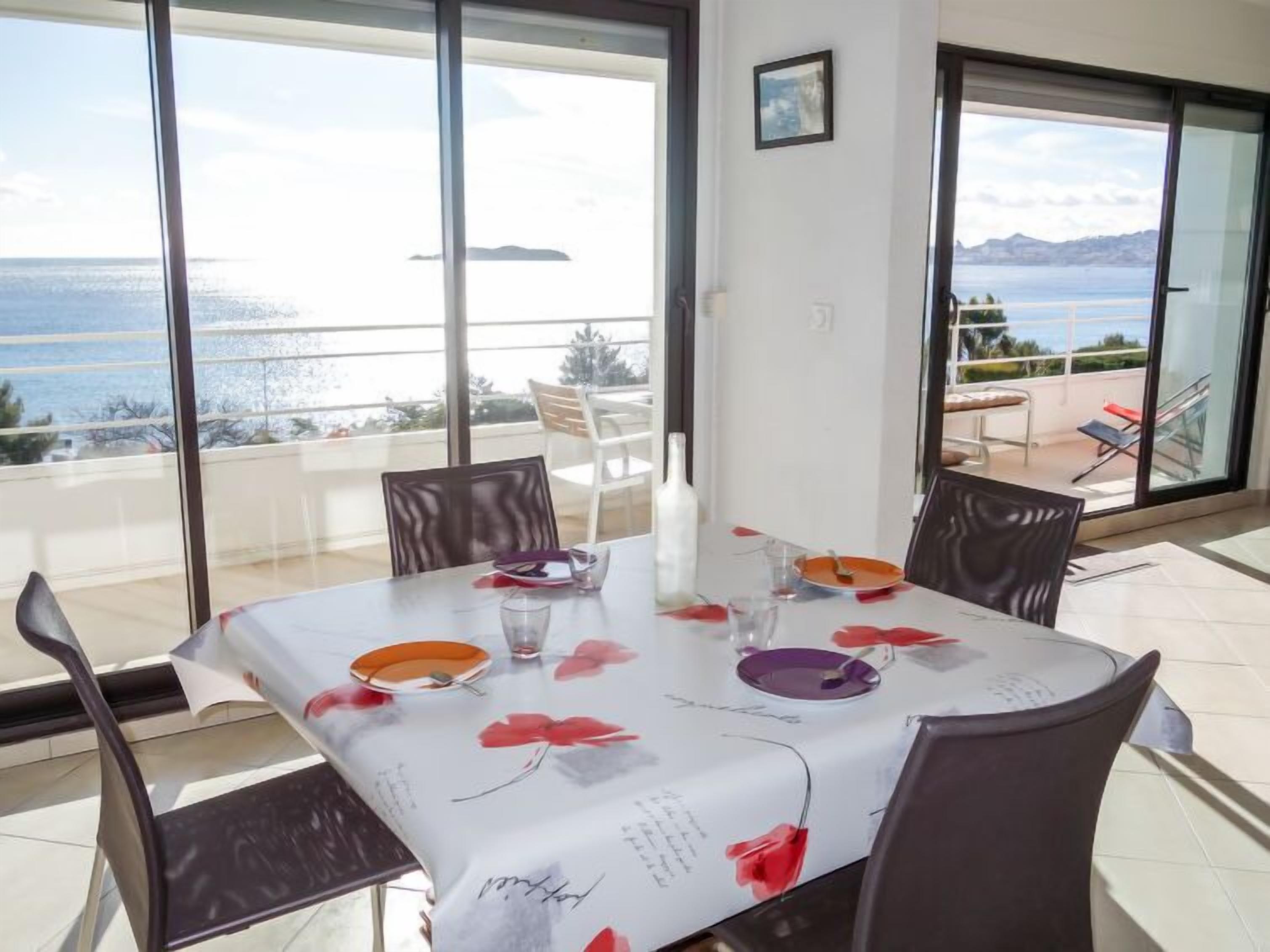 A dining area with a table set for four, large windows offering a view of the sea and mountains, and a balcony with outdoor seating.