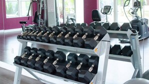 Un gimnasio con estanterías llenas de mancuernas, máquinas de ejercicio y amplias ventanas que dejan entrar luz natural.