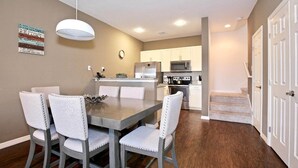 Una cocina moderna con armarios blancos, un refrigerador de acero inoxidable y un microondas. Hay una mesa de comedor con cuatro sillas y una escalera que sube a otro nivel.