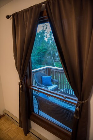 Une fenêtre avec des rideaux marron, donnant sur une terrasse en bois où se trouve une chaise en osier et un coussin bleu.