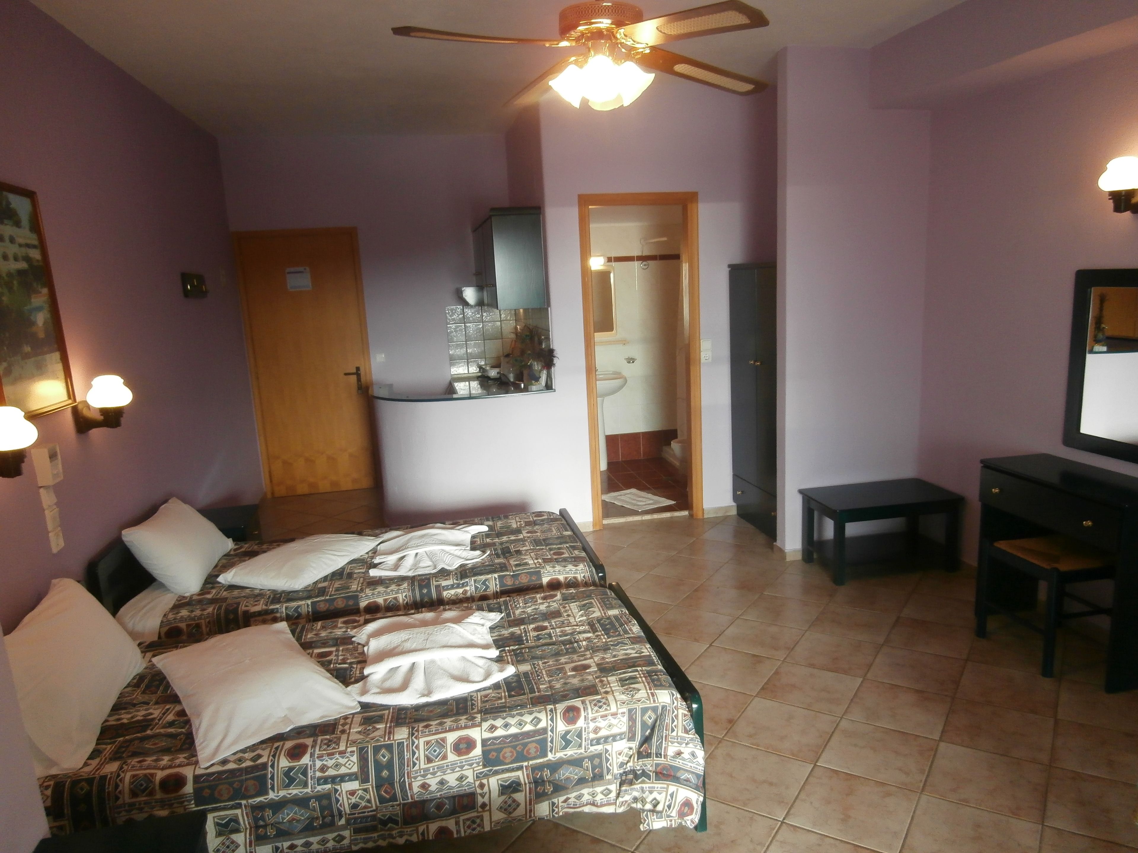 A bedroom with two beds, a desk, a chair, a ceiling fan, and a bathroom visible through an open door.