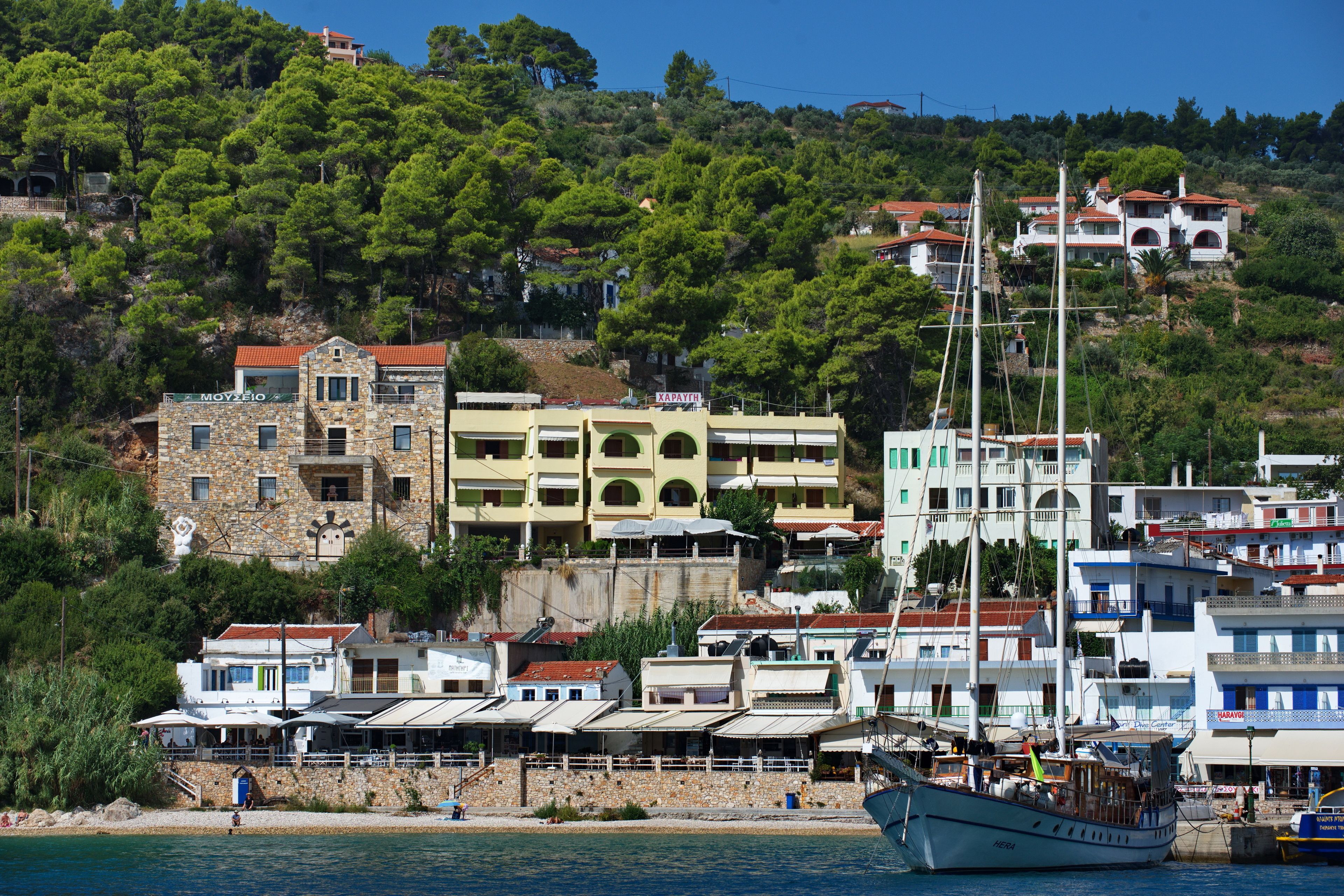 Un pueblo costero con un velero blanco destacado amarrado cerca de la orilla, rodeado de edificios con diversos estilos arquitectónicos, incluyendo construcciones de piedra y modernas.
