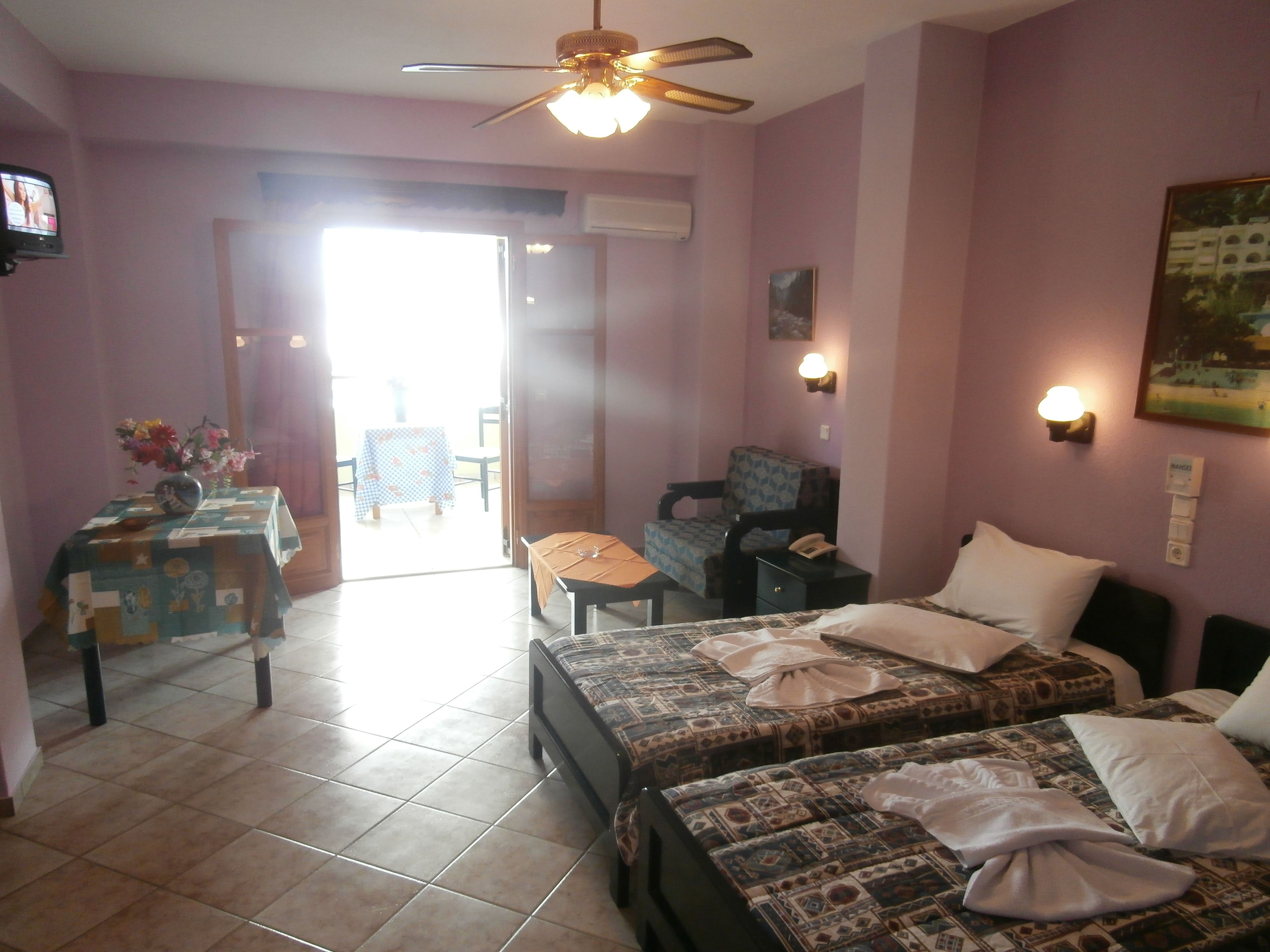 A hotel room with two beds, a desk, a chair, a ceiling fan, and a television.