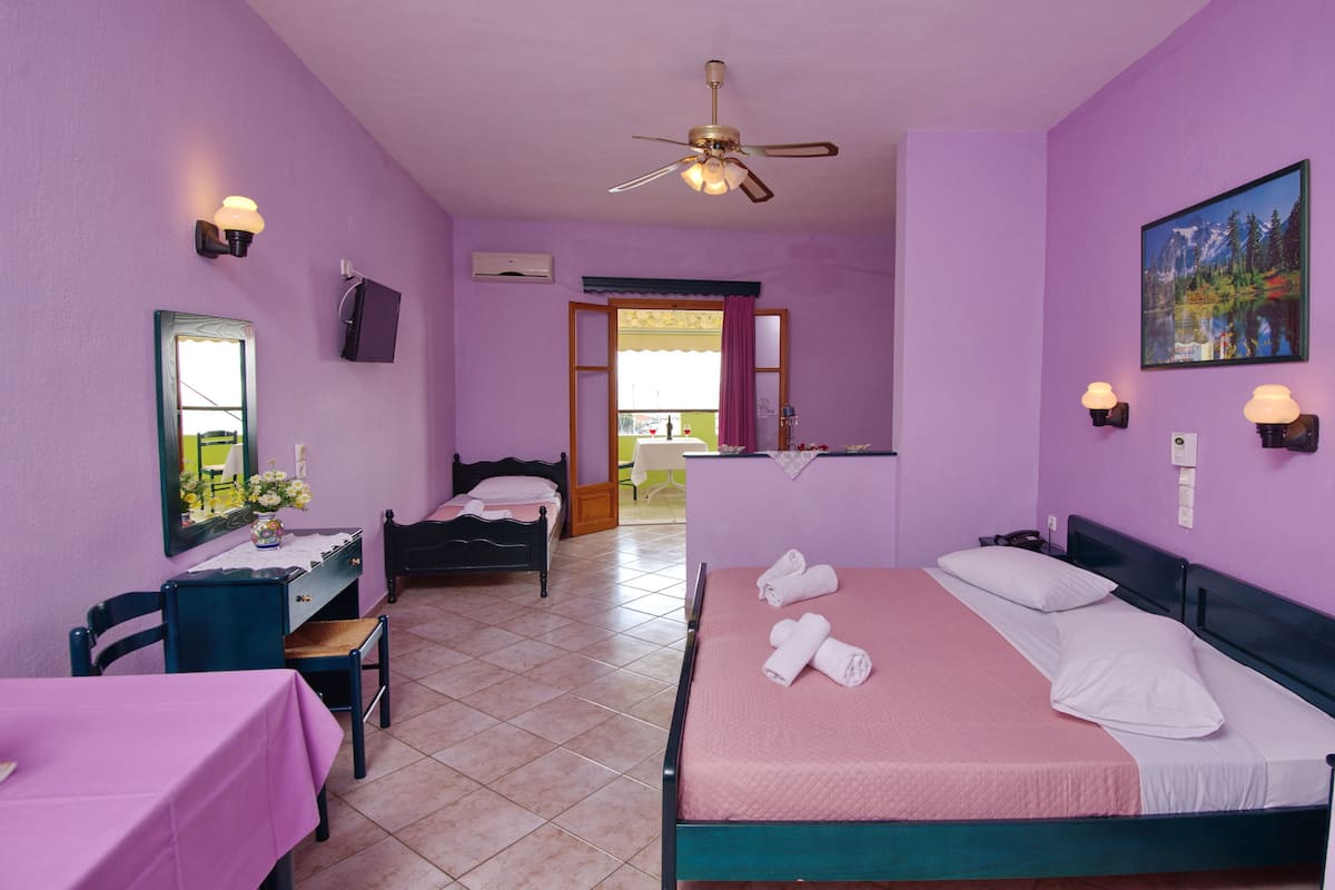 A bedroom with a bed, a desk, a chair, a table, a ceiling fan, and a painting on the wall.