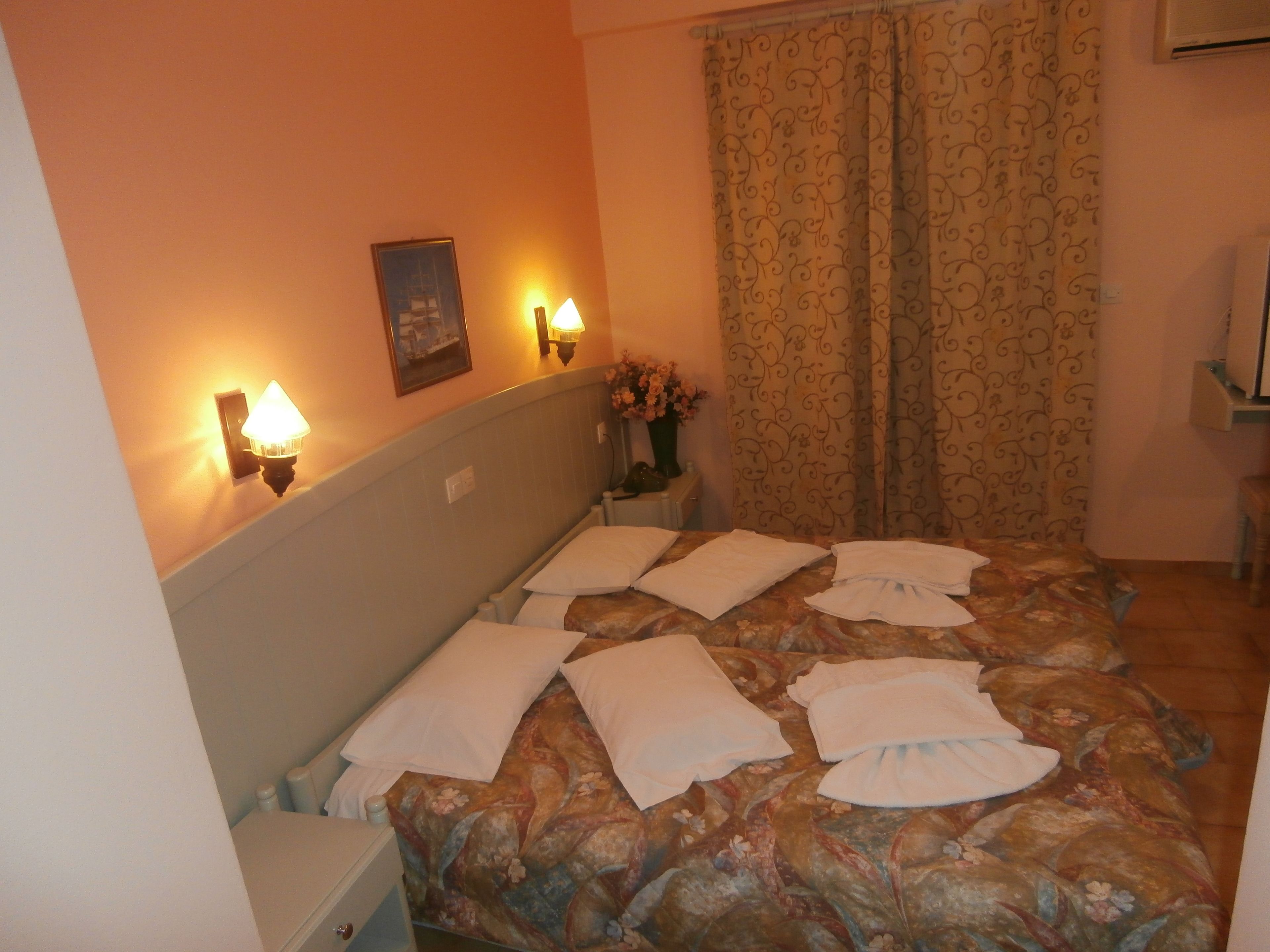 A hotel room with a bed, two bedside lamps, a nightstand, a vase with flowers, and a window with curtains.