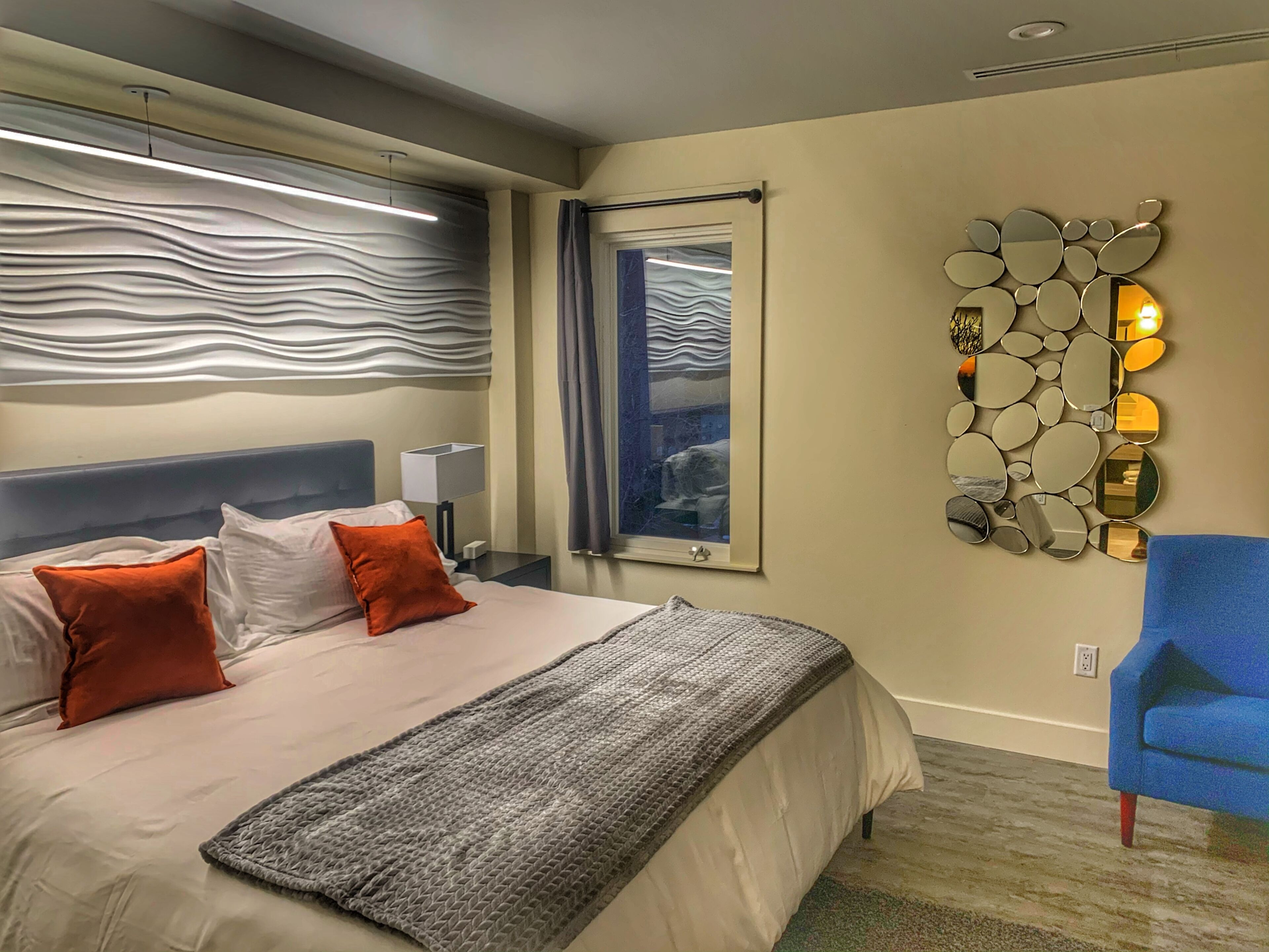 A hotel room with a bed, a blue armchair, a decorative wall piece, and a window with curtains.