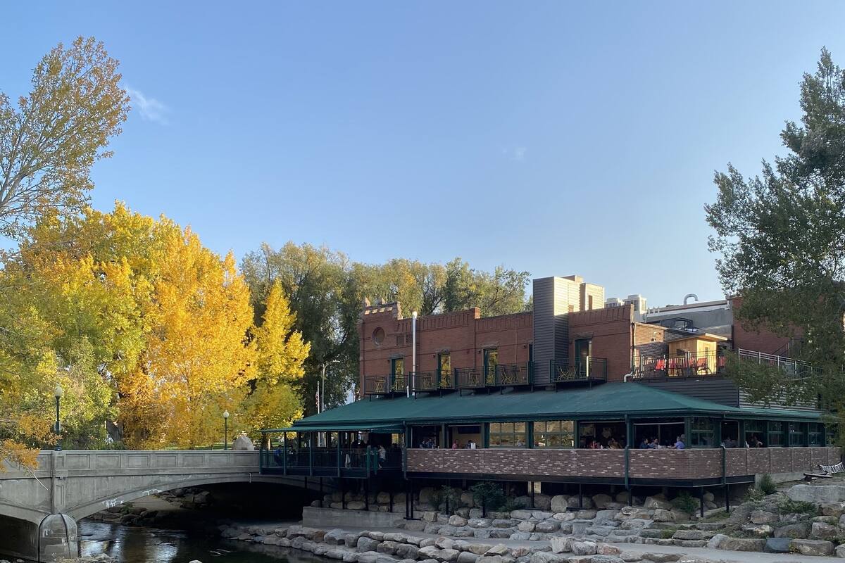 Un restaurante a la orilla del río con terraza, un puente de piedra y follaje otoñal.