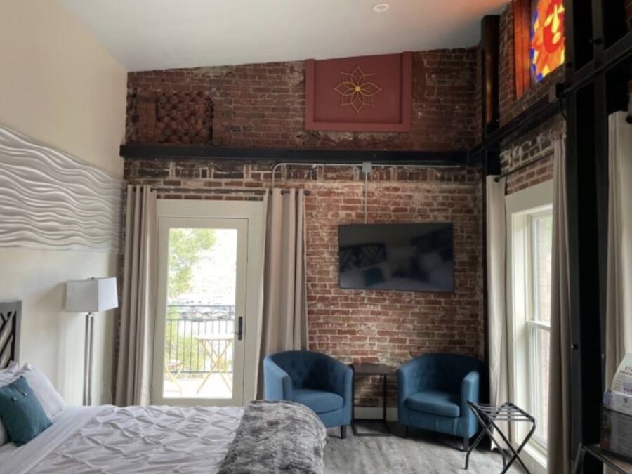 Habitación de hotel con pared de ladrillo, una cama, dos sillones azules, una mesita y un televisor en la pared.
